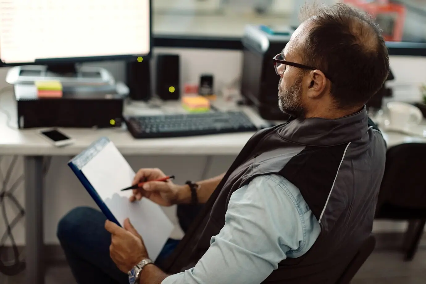 Vorarbeiter nutzt einen Desktop-PC und schreibt Notizen, während er im Büro einer Autowerkstatt arbeitet.